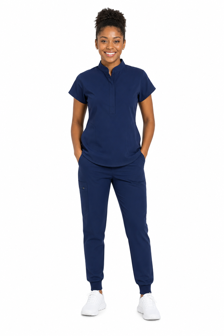 A woman modeling a navy blue scrub set consisting of a short-sleeve mock-neck top and matching jogger pants, paired with white sneakers, against a plain white background.