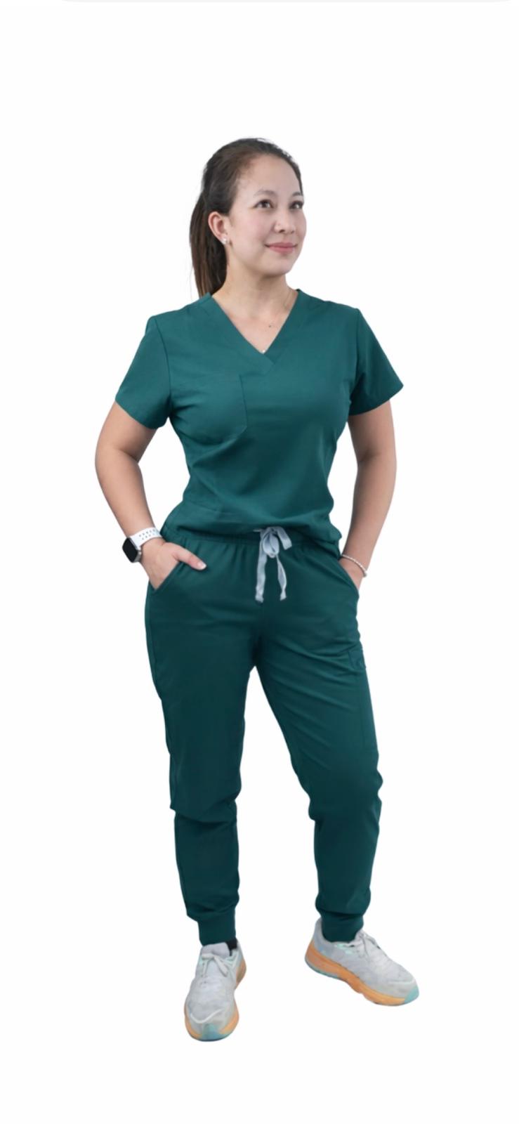 A woman modeling a matching dark green scrub set consisting of a V-neck short-sleeve top and jogger-style pants with a drawstring waist, paired with white sneakers, against a plain white background.